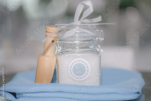 Beautiful Sea Salt filled clear jar with white bow wedding favor with wood on blue blanket in front of white bokeh photo