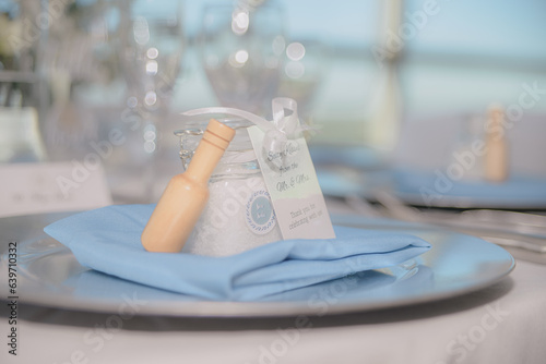 Beautiful Sea Salt filled clear jar with white bow wedding favor with wood on blue blanket on trey in front of white bokeh photo