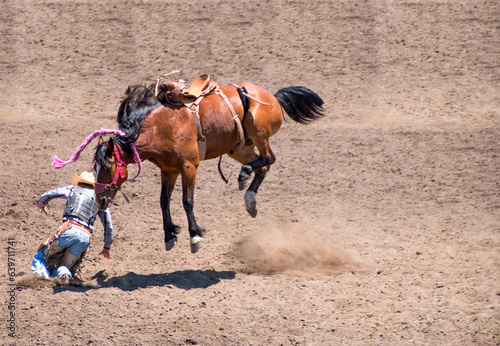 A cowboy has been bucked off a bucking bronco is in front of the horse. He is at a rodeo in an dirt arena. The horse has 4 legs off the ground. There is a red metal gate behind. 