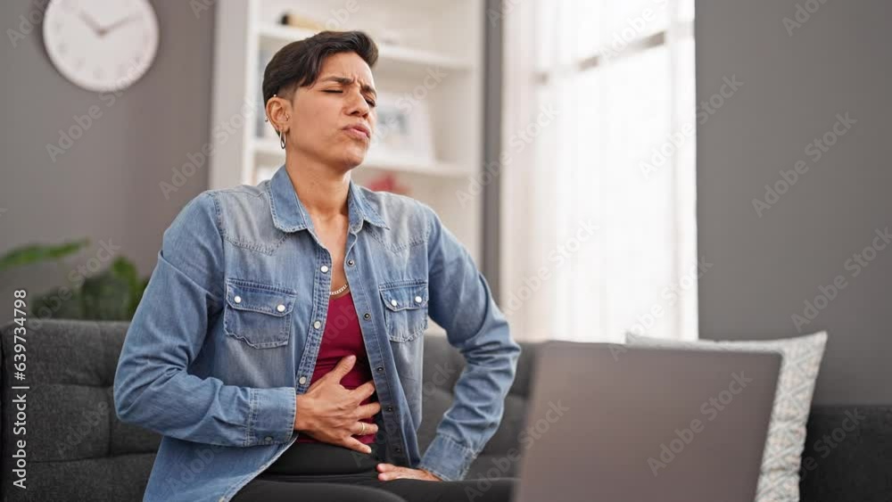 Young beautiful hispanic woman using laptop sitting on sofa suffering for stomachache at home Young beautiful hispanic woman using laptop sitting on sofa suffering for stomachache at home