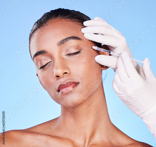 Needle, woman and face in studio for skincare, beauty process and aesthetic filler on blue background. Indian model, hands of surgeon and injection for plastic surgery, facial change or prp cosmetics photo