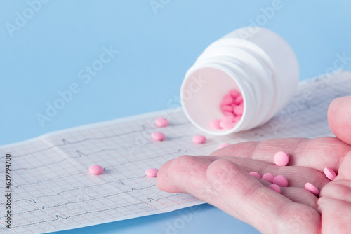 A hand pours a handful of pink pills from a white jar onto an electrocardiogram of the heart, on a blue background. The concept of a healthy lifestyle and timely medical examination.