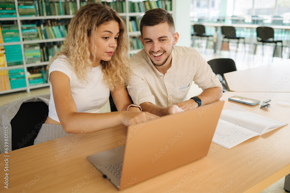 Two classmate look into laptop and laugh. Boy and girl study in library. University concept