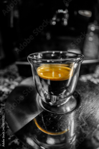 Glass cup with freshly brewed black coffee with foam and golden cream with visible bubbles on top of a metal coffee plate with its reflection of the coffee foam. Dark black and white background.