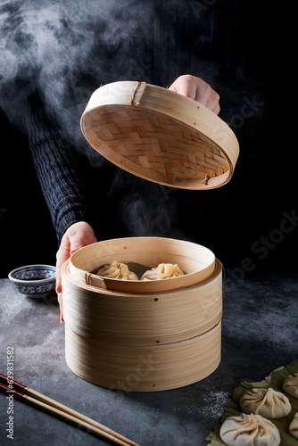 Dim sum bamboo steamer opened. There is steam and freshly made dim sum visible. Hand lifting the top of the dim sum basket. Some raw dim sums on the table.   photo