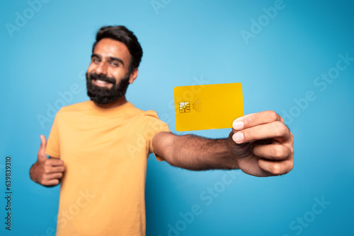Pleased middle aged indian man holding yellow credit card and showing thumb up, selective focus