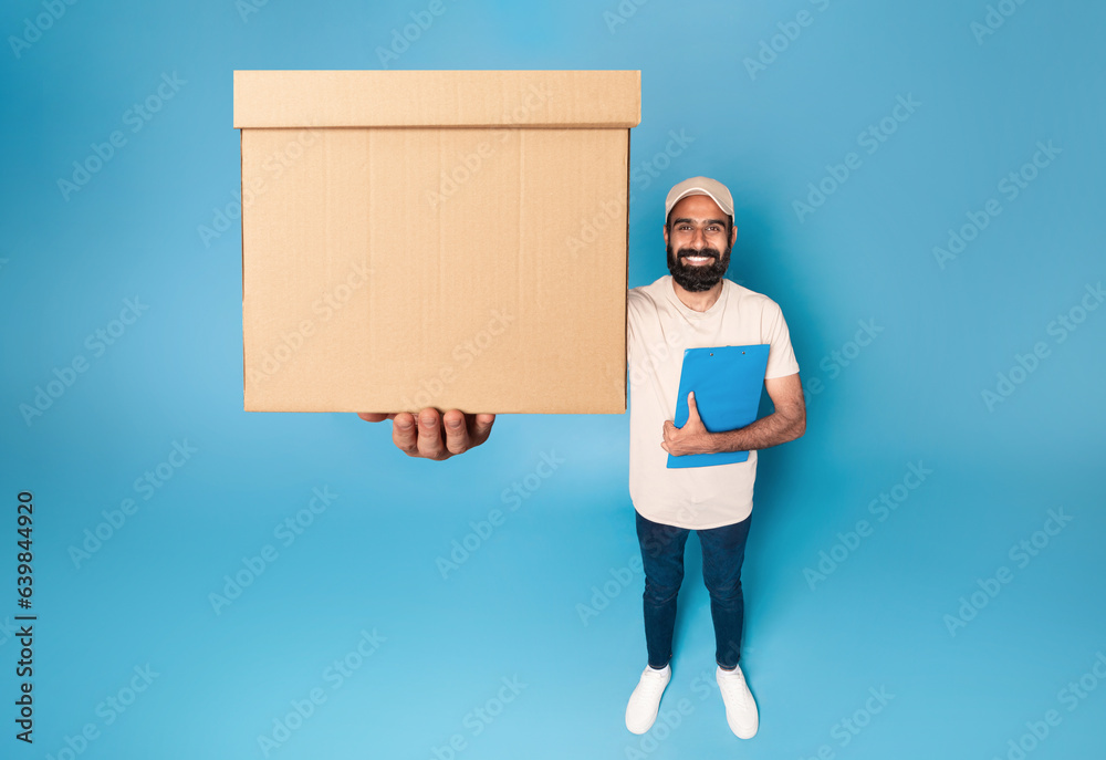 Indian delivery man holding parcel cardboard box stretching to camera, standing over blue background and smiling Indian delivery man holding parcel cardboard box stretching to camera, standing over blue background and smiling