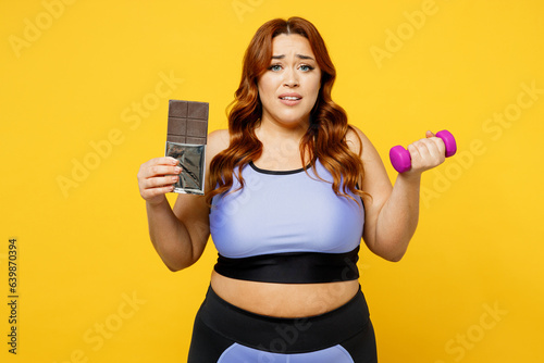 Young sad chubby overweight plus size big fat fit woman wearing blue top warm up training hold dumbbell eat bar of chocolate isolated on plain yellow background studio home gym. Workout sport concept.