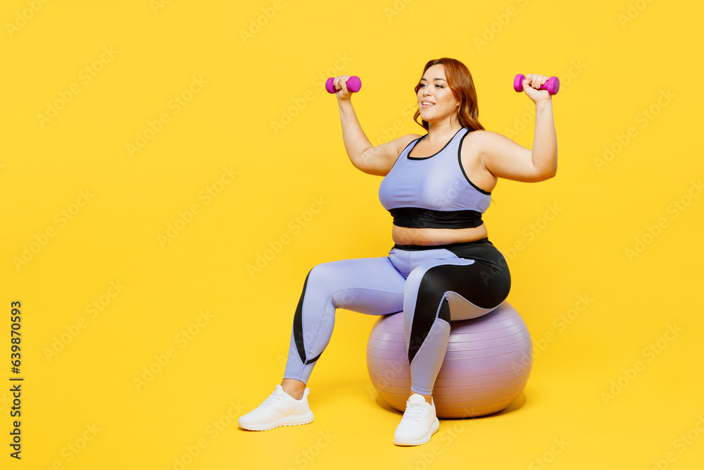 Full body young chubby plus size big fat fit woman wear blue top warm up train sit on fit ball hold dumbbells raise up hands isolated on plain yellow background studio home gym. Workout sport concept. Full body young chubby plus size big fat fit woman wear blue top warm up train sit on fit ball hold dumbbells raise up hands isolated on plain yellow background studio home gym. Workout sport concept.