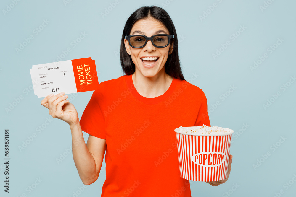 Young surprised fun latin woman in 3d glasses wear orange red t-shirt casual clothes watch movie film hold in hand ticket bucket of popcorn in cinema isolated on plain blue background studio portrait. Young surprised fun latin woman in 3d glasses wear orange red t-shirt casual clothes watch movie film hold in hand ticket bucket of popcorn in cinema isolated on plain blue background studio portrait.