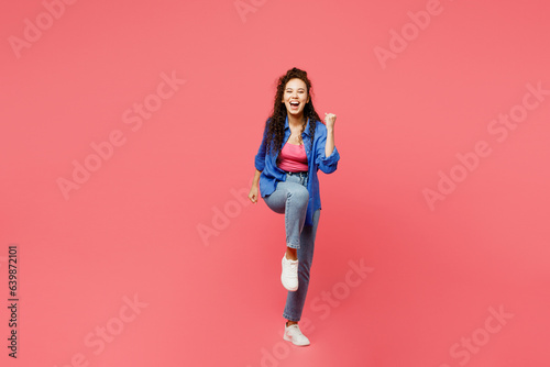 Full body young woman of African American ethnicity wear blue shirt casual clothes doing winner gesture celebrate clenching fists say yes raise up leg isolated on plain pastel pink background studio.