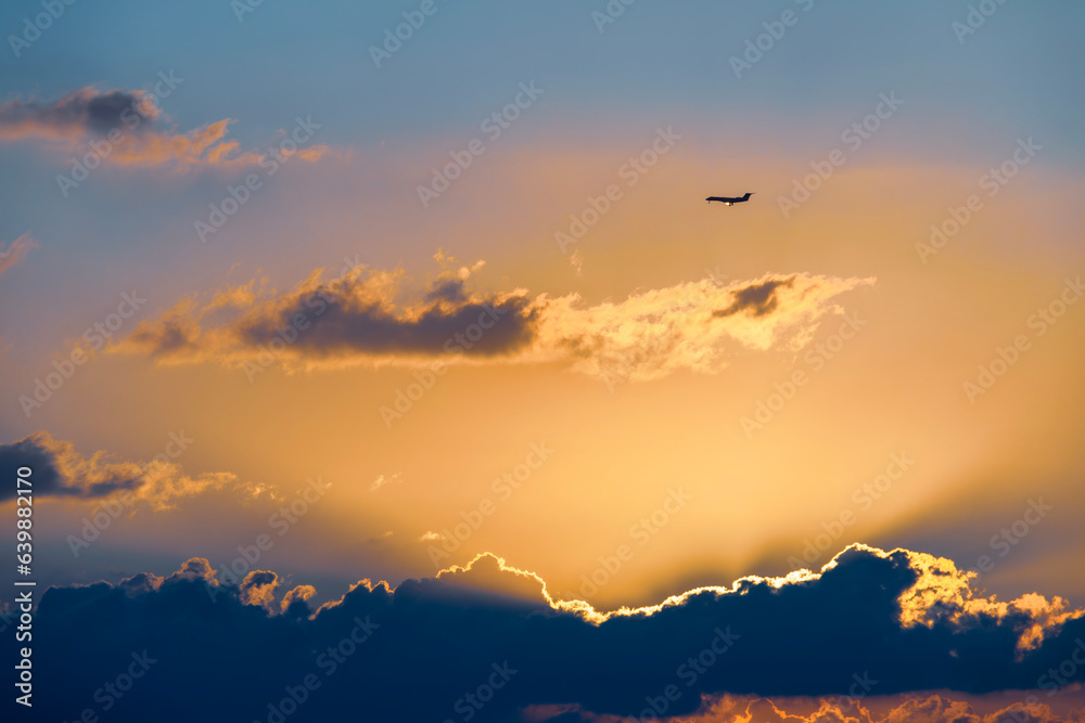 A plane flying in the sky at sunset. The plane is silhouetted against the fiery sky. The sun is setting in the distance, and its rays are visible through the clouds.