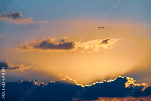A plane flying in the sky at sunset. The plane is silhouetted against the fiery sky. The sun is setting in the distance, and its rays are visible through the clouds.