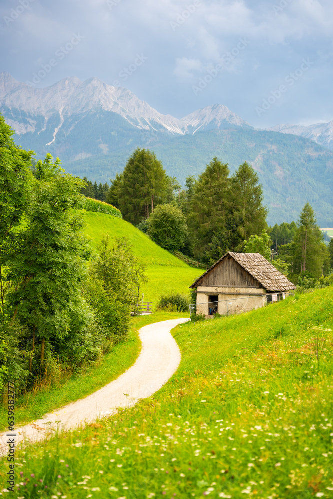 Green meadows with rural road, wooden house, mountains and forest on backgound at sunset in summer, Italy