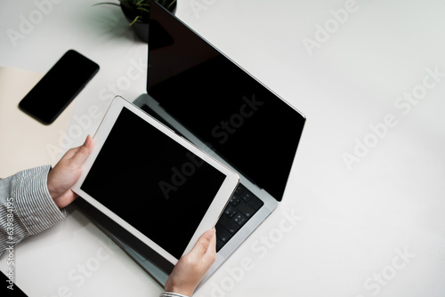 Two hands holding black screen touch pad screen for mockup multimedia technology device with tablet computer and plant on table indoor workspace working on reviewing and reading application document