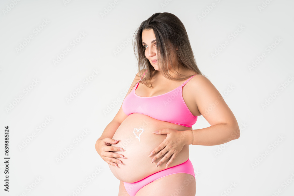 Happy plus size pregnant woman in pink underwear applying skin stretch mark cream on her belly on gray background, maternity clothes, pregnancy concept Happy plus size pregnant woman in pink underwear applying skin stretch mark cream on her belly on gray background, maternity clothes, pregnancy concept