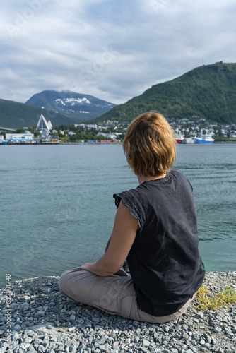 Woman enjoying a view of the Tromso harbor with Arctic Cathedral, fisherman boats and snow covered mountains on the background. Norway 2023