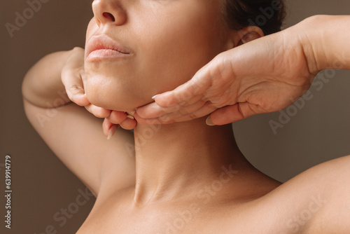 Cropped shot of young caucasian woman touching under the chin with hands massaging her face on dark brown background. Rejuvenation, facelift, facefitness. Exercises from the second chin, pelican neck photo