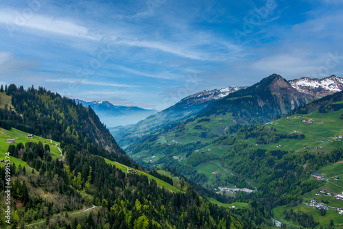 Spring sunlight illuminating the forests, farms, villages and the partially snow-covered mountains of the Großes Walsertal in the Austrian Alps, with mist covering the valley in the background