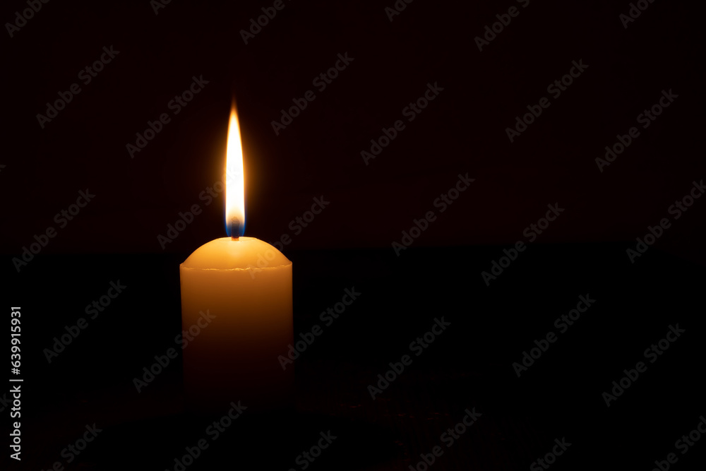 Single burning candle flame or light glowing on a big white candle on black or dark background on table in church for Christmas, funeral or memorial service with copy space. Single burning candle flame or light glowing on a big white candle on black or dark background on table in church for Christmas, funeral or memorial service with copy space.