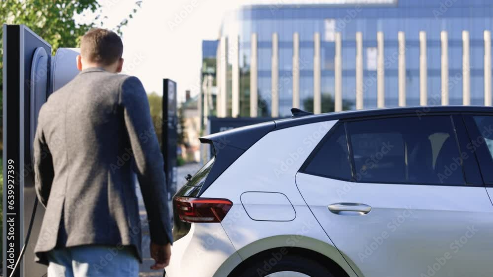 Caucasian businessman attaching power cable to electric car. Electric vehicle recharging battery charging port plugging in EV modern car. Save ecology alternative energy sustainable of future Caucasian businessman attaching power cable to electric car. Electric vehicle recharging battery charging port plugging in EV modern car. Save ecology alternative energy sustainable of future