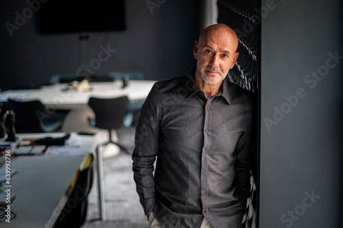 Confident mid aged businessman standing at the wall in a modern office and looking at camera