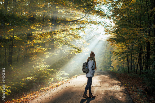 Woman with backpack walks on road in autumn forest with sunbeam shining through trees. Atmospheric mood in cold foggy morning. Hiking in nature