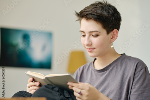 Teenage girl with short dark straight hair looking through text of novel on pages of book in grey cover while sitting in front of camera