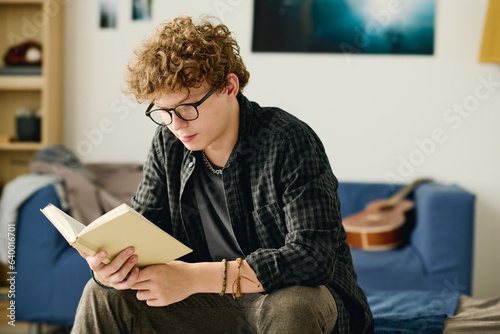 Foto Serious male teenager in eyeglasses reading curious novel while sitting on bed i