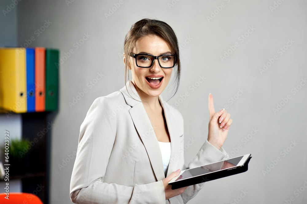 Young excited woman pointing at free copy space on gray wall while holding a tablet, wearing glasses and formal clothes. Mock-up for advertising online courses or workshops. Discount, promotion, sale.