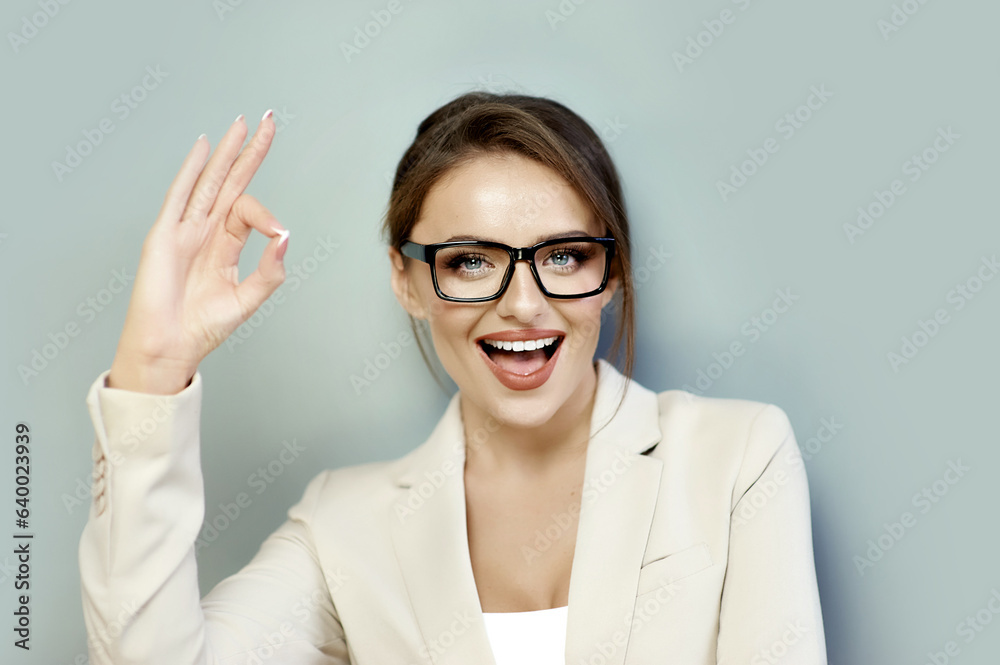 Pretty joyful caucasian woman in black-framed glasses showing OK sign isolated on gray background with copy space. Approval, support, agreement, excellent choice, good job. Pretty joyful caucasian woman in black-framed glasses showing OK sign isolated on gray background with copy space. Approval, support, agreement, excellent choice, good job.