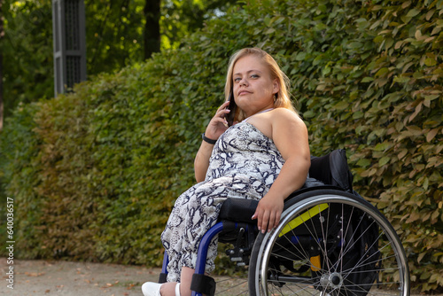 Smiling Young Woman With Short Stature On Wheelchair Talks On Smartphone in Green Park At Summer Day. Female Adult With Disability. Copy Space For Text. Horizontal Plane