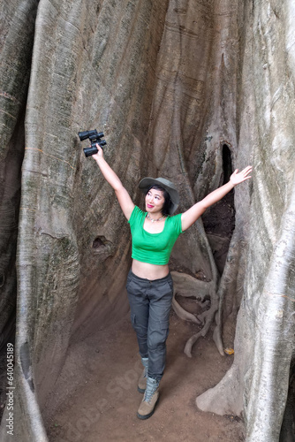  Beautiful Asian woman Holding binoculars at the roots of a large tree and are delighted happily. This tree is very large and is a popular attraction for Thai tourists.