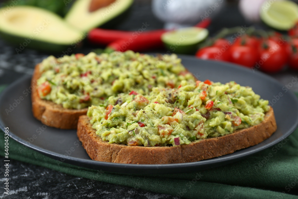 Slices of bread with tasty guacamole and ingredients on black textured table, closeup