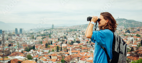 A beautiful girl traveler looks through binoculars at Sarajevo, the capital of Bosnia and Herzegovina. View of the Balkan European city with orange roofs in the mountains from a view point