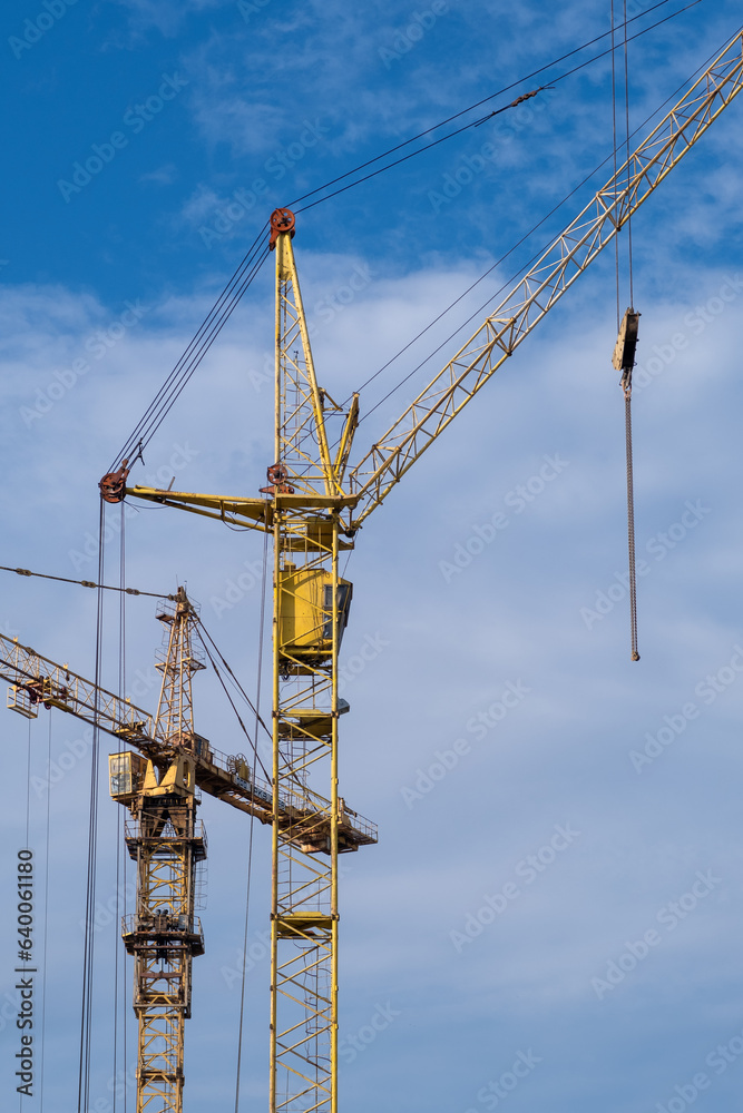A crane and a building under construction against a blue sky background. Builders work on large construction sites, and there are many cranes working in the field of new construction. A crane and a building under construction against a blue sky background. Builders work on large construction sites, and there are many cranes working in the field of new construction.
