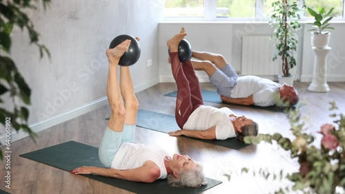 Senior woman in company of elderly friends and daughter perform Pilates exercises with softball. Visitior yoga class lying on back, raised legs perpendicular to floor and clamped softball in feet photo