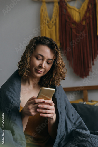 Young woman with smartphone writing text message in bed at home
