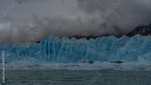 A wall of blue ice with cracks and sharp spikes rises above a turquoise lake. Breakaway melting icebergs float in the water. Mountains in the fog. Perito Moreno glacier. Los Glaciares National Park.