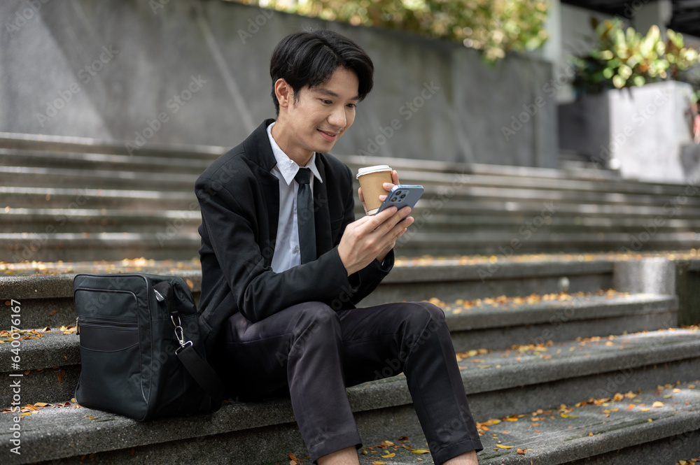 A happy Asian businessman is using his smartphone and sipping coffee while sitting on a staircase A happy Asian businessman is using his smartphone and sipping coffee while sitting on a staircase