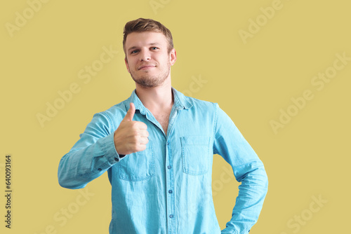 Handsome young caucasian man in denim shirt showing thumbs up on yellow background. Positive confident man. Approving gesture. Looks into camera.