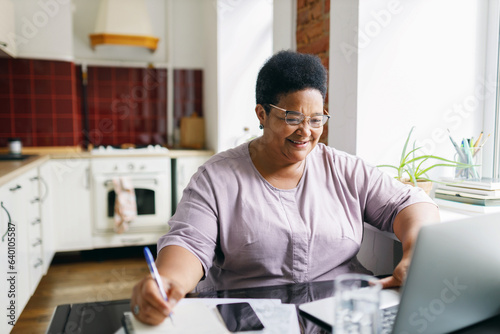 Happy cheerful african american overweight female of 60s using laptop sitting at kitchen table, smiling looking at screen, noting something down, making list of purchases, checking sales