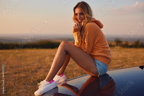 Beautiful young woman in orange hoodie is sitting on the hood of a car and enjoying a during sunset