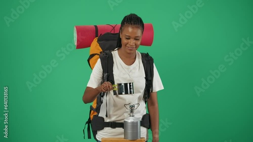 Woman tourist with backpack boils water in a pot on a travel gas burner. Isolated on green background. photo