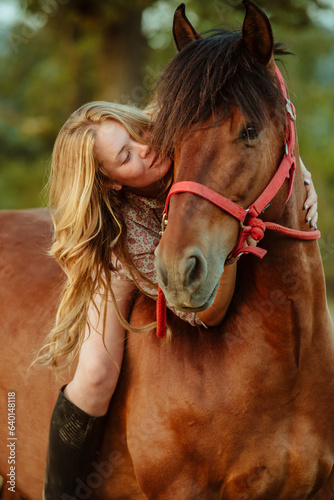 Portrait of a young beautiful cute blond cowgirl is hugging her horse at sunset. Equitation concept.