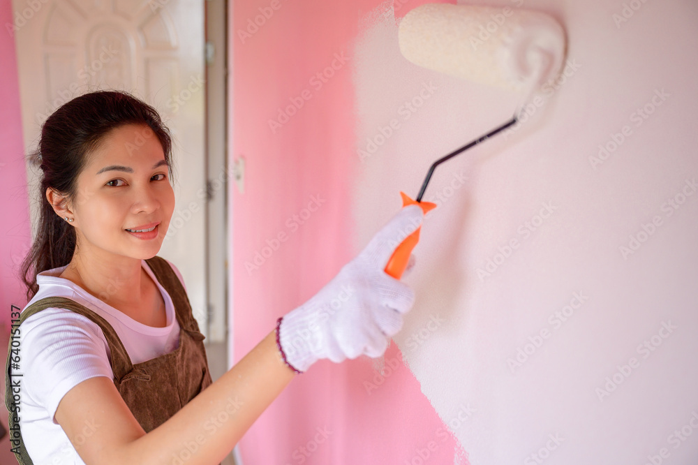 Young Asian woman looking at camera holding paint roller Asian woman painting to change the wall color of her house to white home interior ideas Young Asian woman looking at camera holding paint roller Asian woman painting to change the wall color of her house to white home interior ideas