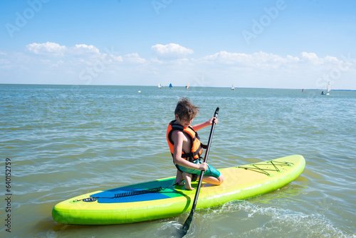 A 10 year old boy is learning to ride a SUP board in the sea. a child in an orange life jacket rides a sapboard alone on the sea on a summer vacation on a sunny day. photo