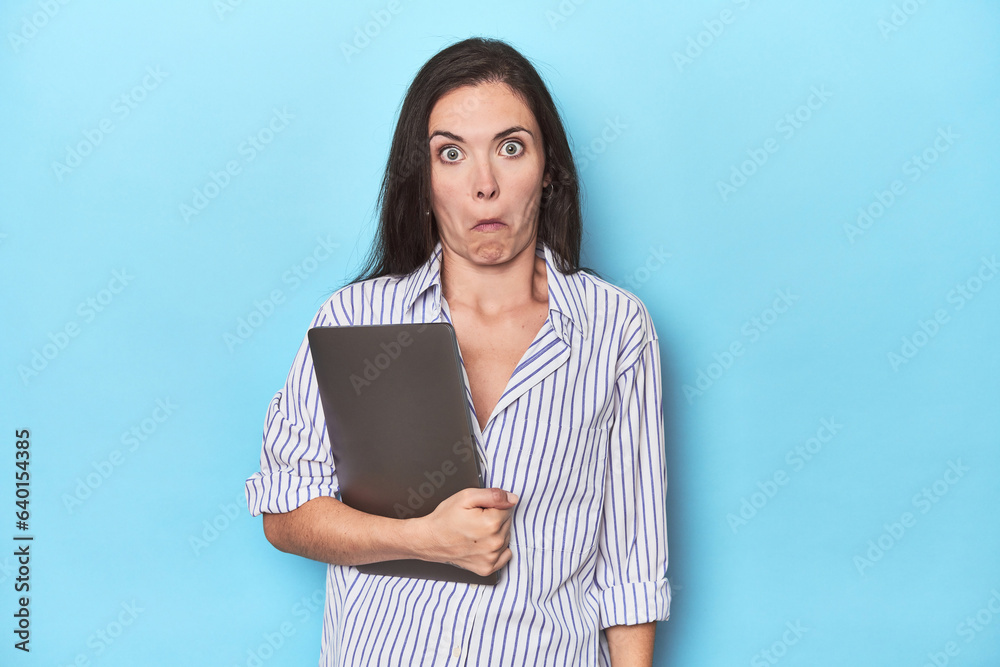 Young businesswoman holding laptop on blue shrugs shoulders and open eyes confused. Young businesswoman holding laptop on blue shrugs shoulders and open eyes confused.