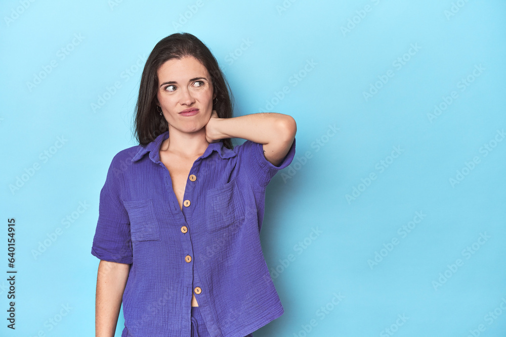 Fototapeta premium Young caucasian woman on blue backdrop touching back of head, thinking and making a choice.