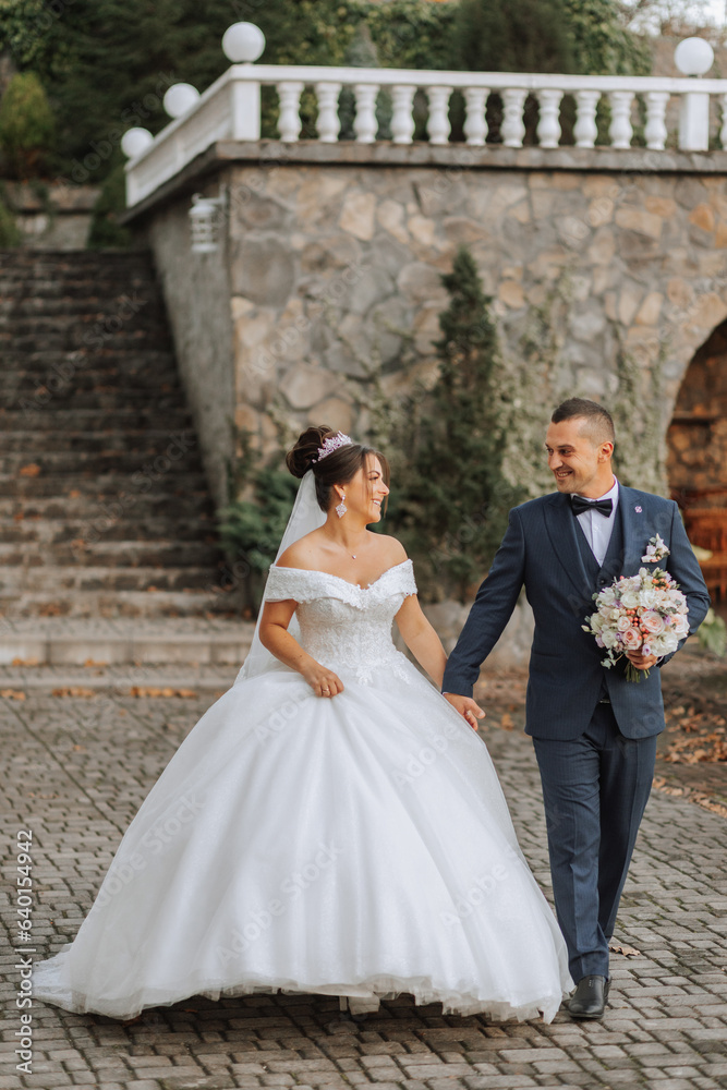 Gorgeous wedding couple walking on stone stairs near old castle in park. Stylish beautiful bride in amazing gown and groom posing on background of ancient building